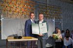 Feierliche Übergabe einer Auszeichnung an eine Person. Im Hintergrund eine Wand mit floraler Dekoration.