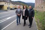 Drei Personen stehen nebeneinander auf einem Gehweg vor Gebäuden und bewaldeten Bergen.