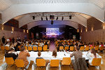 Versammlung in heller Halle. Menschen sitzen an Tischen, Blick auf eine Bühne mit Präsentation im Hintergrund.