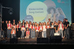 Gruppe junger Menschen steht auf einer Bühne vor Publikum. Im Hintergrund ein Banner mit der Aufschrift 'Kinderrechte Songcontest'.