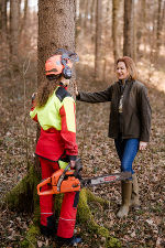 Bildbeschreibung: Eine Person in Schutzkleidung steht mit einer Kettensäge neben einer weiteren Person im Wald.