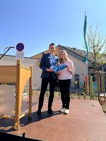 Zwei Personen stehen vor einem Gebäude mit einem gelben Schild und grünen Büschen. Im Hintergrund eine steirische Flagge.