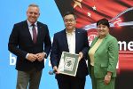 Bildbeschreibung: Zwei Männer und eine Frau stehen vor einer Grafik mit chinesischer Fahne. Ein Mann hält einen Bilderrahmen in der Hand.
