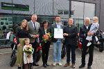 Bildbeschreibung: Gruppe von Menschen bei einer feierlichen Übergabe von einer Urkunde und Blumen vor einem Motorradgeschäft.