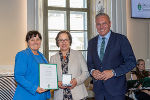 Drei Personen im feierlichen Rahmen. Zwei Personen überreichen eine Urkunde an eine dritte Person.