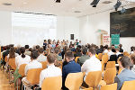 Versammlung in einem hellen Saal. Eine Gruppe junger Menschen steht im Fokus, umgeben von Sitzenden.