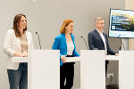 Bildbeschreibung: Drei Personen stehen vor Rednerpulten.