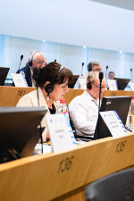 Bildbeschreibung: Eine Frau mit Kopfhörern sitzt in einem Plenum.