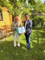 Bildbeschreibung: Festliche Übergabe im Grünen: Eine Person überreicht einer anderen eine Urkunde. Sonnendurchflutete Szene.