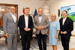 Gruppe von fünf Personen steht in einem Innenraum vor einer Wand mit Fahnen und einem Bildschirm.