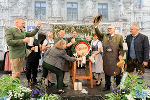 Gruppenfoto bei einem öffentlichen Event. Menschen in Tracht zapfen Bier von einem Fass. Im Hintergrund ein Gebäude mit Säulen und Dekorationen.