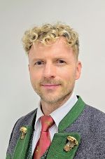 Porträt einer Person mit blonden Locken in traditioneller Tracht mit grauer Jacke und roter Krawatte.