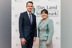 Zwei Personen in formeller Kleidung stehen lächelnd vor einer Wand mit dem Steiermark-Logo.